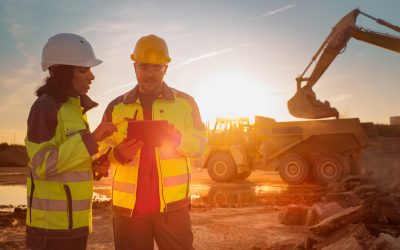 Caucasian Male Engineer Using Tablet And Talking To Hispanic Female Architect On Construction Site Of Real Estate Project. Colleagues Discussing Building Plan Of Apartment Complex During Golden Hour.