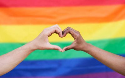 Heart shape made out of two diverse ethnicity hands with a background of a gay pride flag. White and black hands.